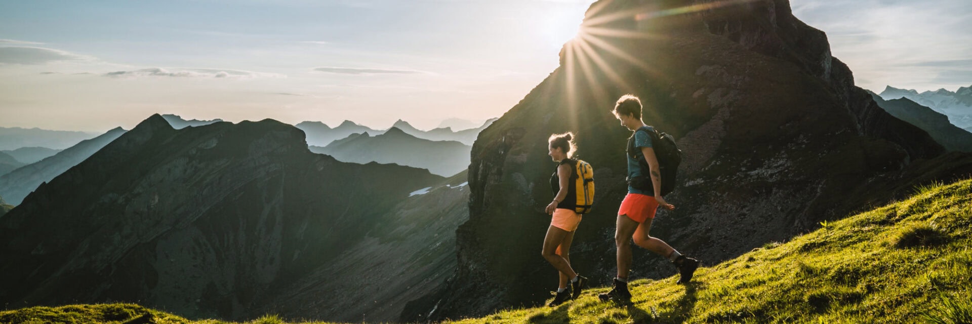 Zwei Wanderer mit Rucksäcken gehen auf einem schmalen Bergpfad durch eine grüne Alpenlandschaft. Im Hintergrund ragen schroffe Gipfel auf, während die tief stehende Sonne hinter einem Berg hervorstrahlt und die Szene in warmes Licht taucht