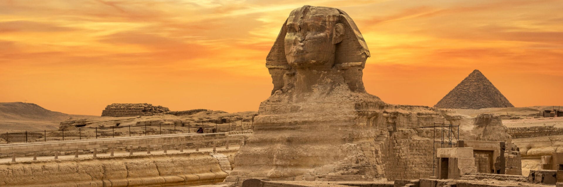 Die Große Sphinx von Gizeh im Vordergrund, aus Kalkstein gemeißelt, mit einer Pyramide im Hintergrund bei warmem orangefarbenem Sonnenuntergang über der Wüstenlandschaft Ägyptens.