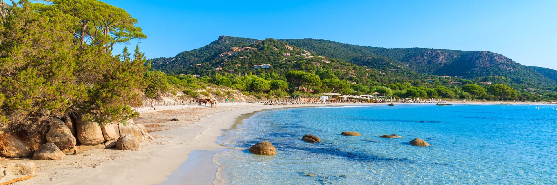 Sonniger Sandstrand mit türkisblauem, klarem Meer, flachen Felsen im Wasser und grünen Pinien; im Hintergrund bewaldete Hügel mit vereinzelten Häusern und einer Strandpromenade mit Liegen.