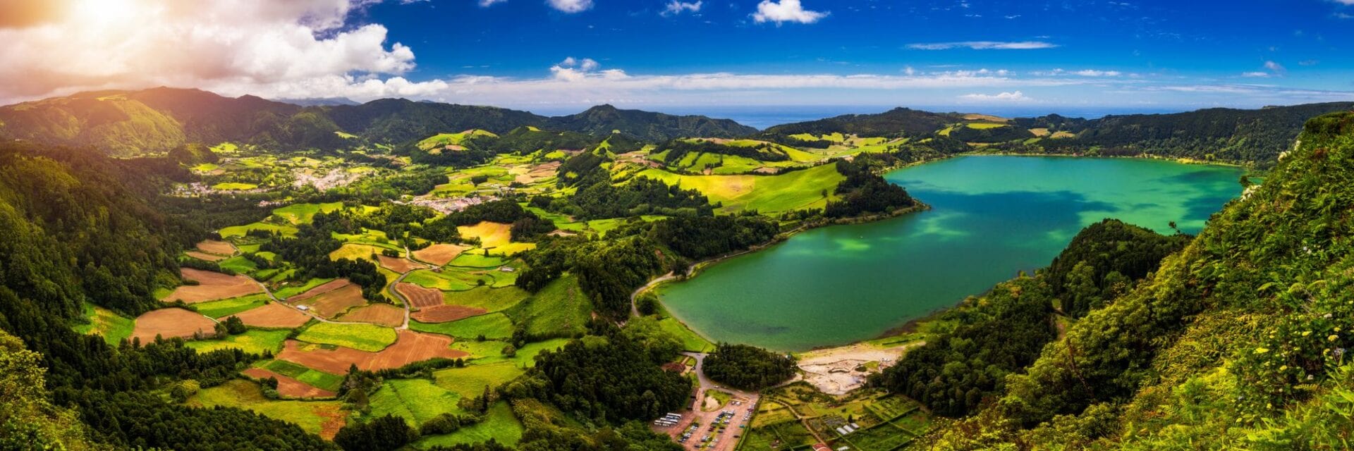 Panoramablick auf die grüne Landschaft von Furnas auf São Miguel mit Feldern, Hügeln und einem türkisfarbenen Kratersee unter blauem Himmel.