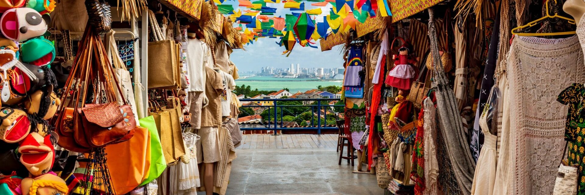 Bunter Souvenirmarkt mit Taschen, Textilien und Handwerkskunst; über dem Gang hängen farbige Fahnen, im Hintergrund Blick auf Meer und Skyline einer brasilianischen Küstenstadt.