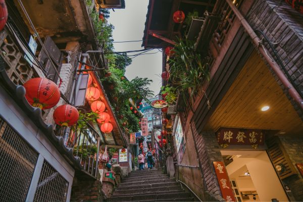 Enge, steile Gasse in Taipeh mit Treppenstufen, traditionellen Gebäuden und zahlreichen roten Lampions, die zwischen Häusern hängen; kleine Geschäfte und üppiges Grün säumen den Weg.