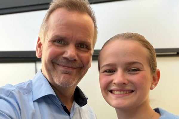 Zwei Personen lächeln in die Kamera: Links ein Mann mittleren Alters mit kurzem Haar und hellblauem Hemd, rechts eine junge Frau mit zusammengebundenen Haaren. Beide stehen nah beieinander in einem hellen Innenraum