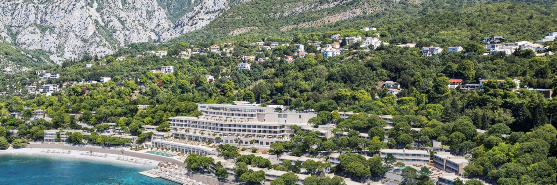 Luftaufnahme eines modernen Strandhotels an der Adriaküste, umgeben von sattem Grün, Pinienbäumen und weißen Villen, mit Bergen im Hintergrund und türkisblauem Meer im Vordergrund.