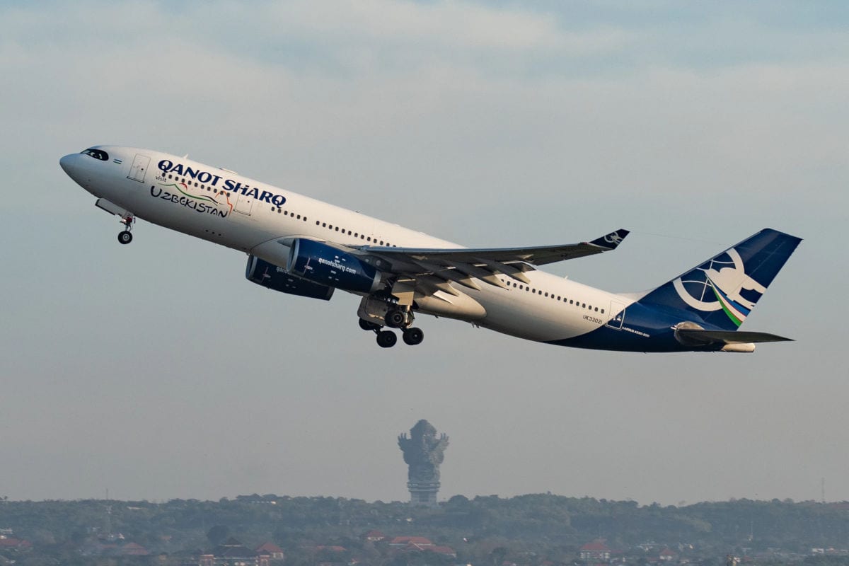 Ein modernes Passagierflugzeug der Fluggesellschaft Qanot Sharq hebt mit ausgefahrenem Fahrwerk vom Flughafen ab, deutlich erkenntlich an der Beschriftung „Uzbekistan“ und dem blauen Leitwerk. Im Hintergrund ist eine grüne Landschaft mit vereinzelten Gebäuden und einer auffälligen Statue zu sehen.