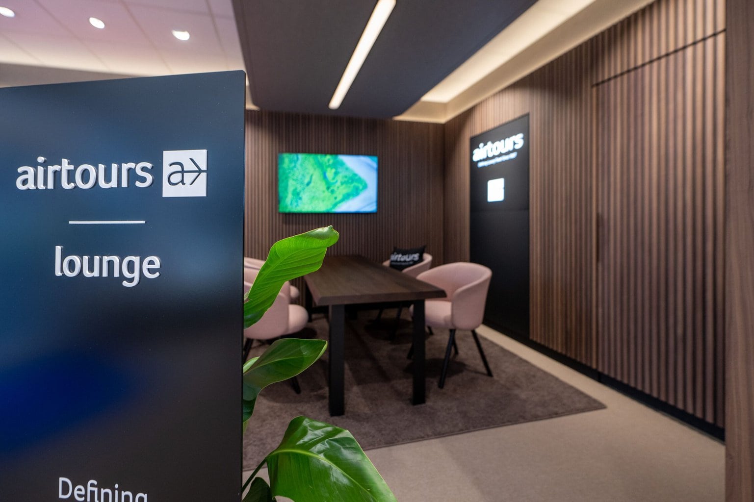 Blick in die Airtours Lounge mit Beratungstisch, Stühlen, Bildschirm und markanter Holzvertäfelung.