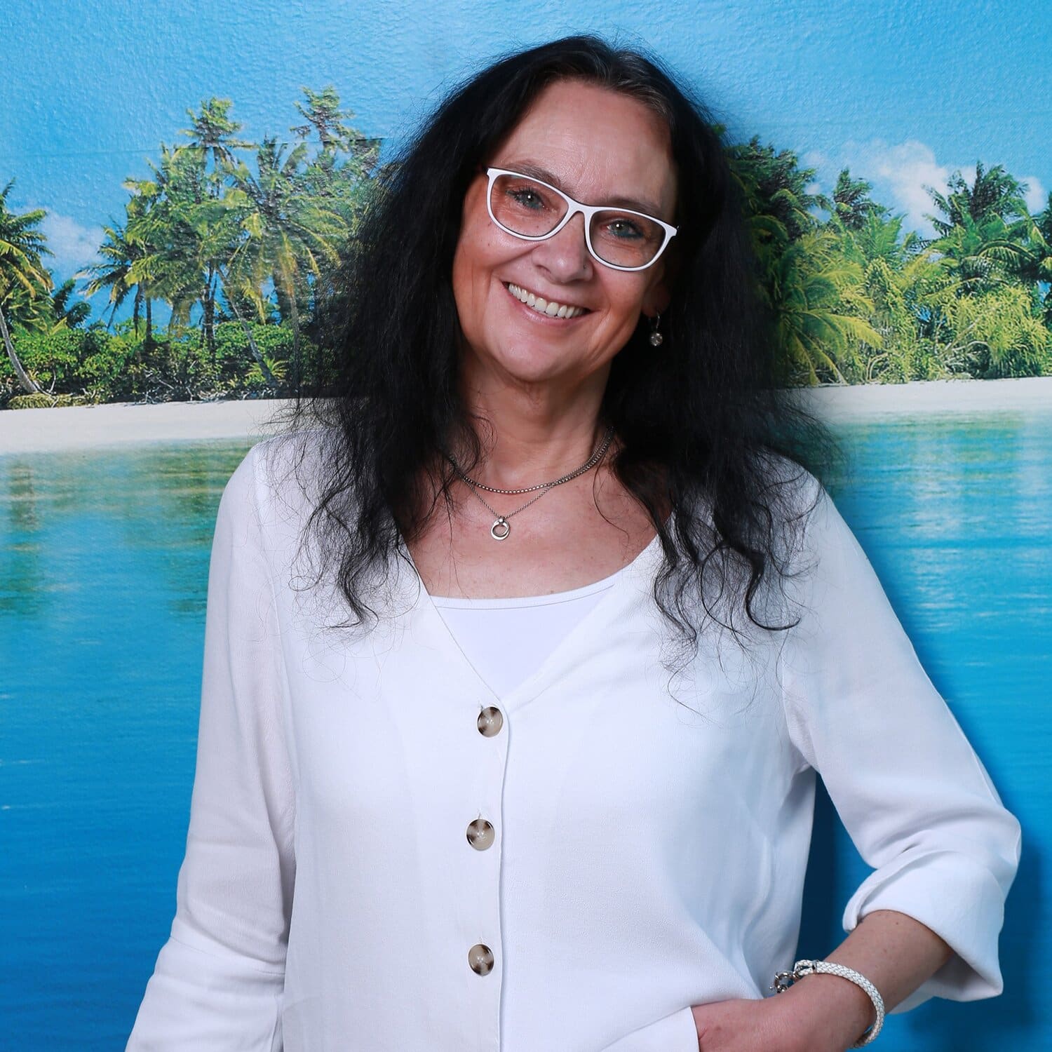 Eine Frau mit weißer Brille und langen schwarzen Haaren lehnt an der Wand vor einer Fototapete mit Palmen und Meer.
