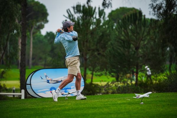Ein Golfer im hellblauen Langarmshirt und beiger Shorts schlägt auf einem grünen Fairway ab, im Hintergrund stehen Bäume und ein Werbebanner von SunExpress mit einem Flugzeugmotiv.