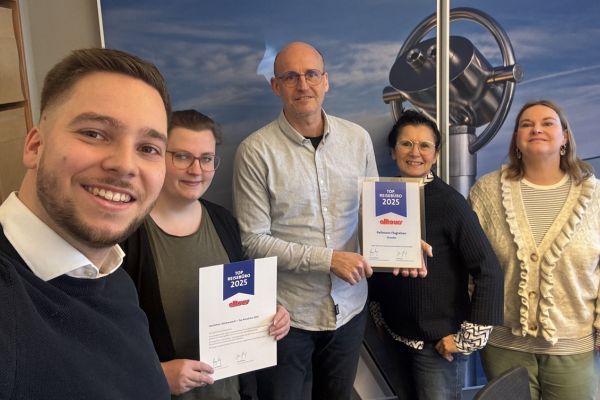 Fünf Personen stehen in einem Büro vor einer Wand mit großem Bildmotiv und präsentieren zwei Urkunden der Auszeichnung „Top Reisebüro 2025“ von Alltours.