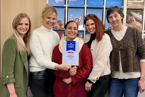 Fünf Frauen stehen in einem Reisebüro vor einer Wand mit Reisemotiven und präsentieren gemeinsam eine Urkunde „Top Reisebüro 2025“ von Alltours.