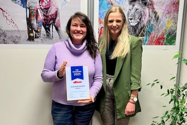Zwei Frauen stehen in einem Büro vor gerahmten Bildern und halten eine Urkunde der Auszeichnung „Top Reisebüro 2025“ von Alltours.