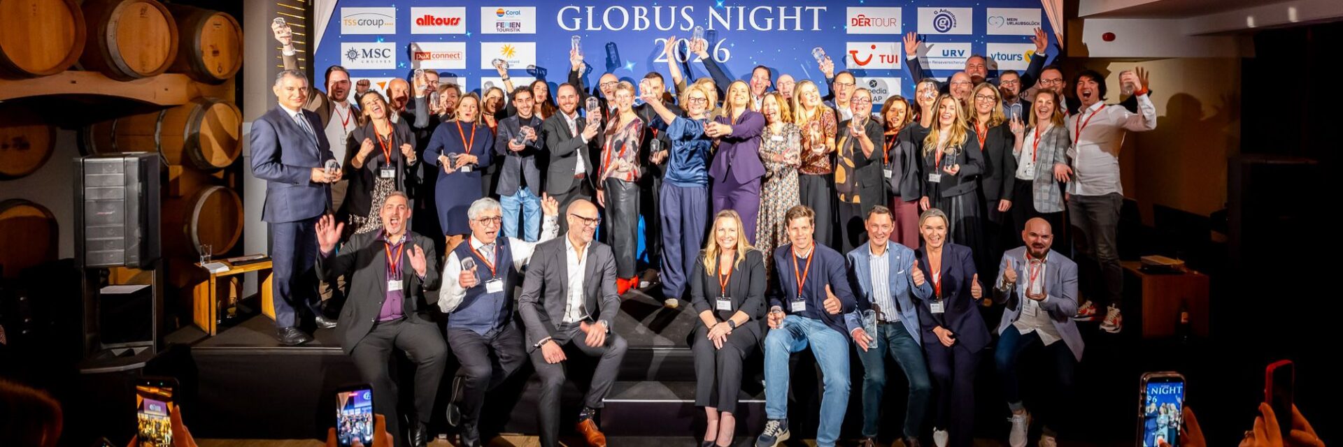 Große Gruppe von festlich gekleideten Erwachsenen auf einer Bühne, die lachend Glastrophäen in die Höhe halten. Im Hintergrund ein Veranstaltungsbanner mit der Aufschrift „Globus Night 2026“ und zahlreichen Unternehmenslogos; die Stimmung ist feierlich und ausgelassen.