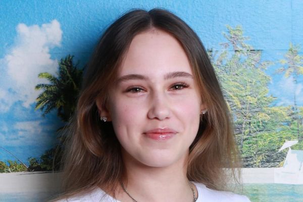 Eine junge Frau mit langen, hellbraunen Haaren steht vor einem tropischen Hintergrund mit Palmen, blauem Himmel und weißen Wolken. Das Foto zeigt sie von vorne, sie trägt ein weißes Oberteil und dezenten Schmuck