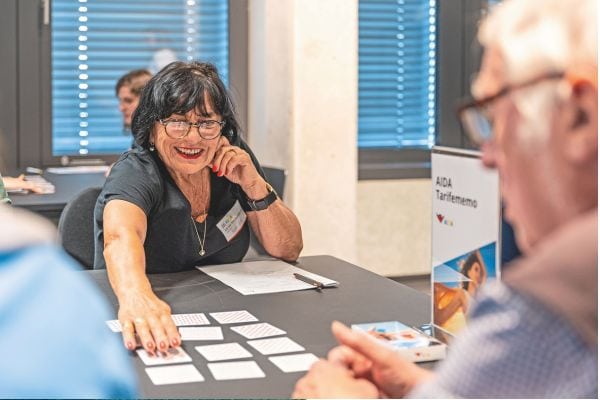 Eine lächelnde Frau sitzt an einem Tisch und spielt mit zwei älteren Personen ein Kartenspiel in einem hellen Seminarraum.