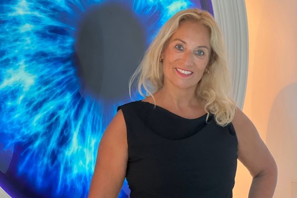 Auf dem Bild ist eine Frau in einem eleganten schwarzen Kleid zu sehen, die vor einer großen, künstlerisch dargestellten blauen Iris steht