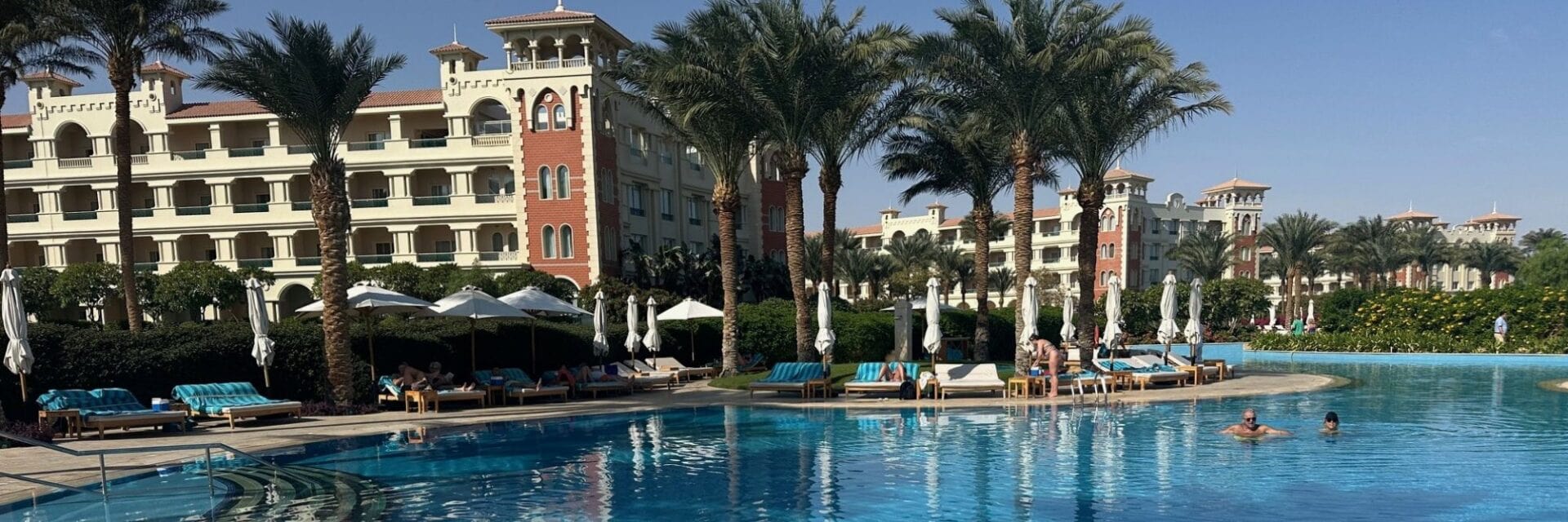 Großer Hotelpool in einer luxuriösen Resortanlage mit Palmen, Sonnenliegen und Sonnenschirmen; im Hintergrund mehrstöckige Hotelgebäude im mediterranen Stil, einige Gäste entspannen am Pool oder schwimmen im Wasser.