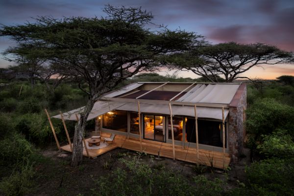 Auf dem Bild ist eine luxuriöse Safari-Lodge in der Serengeti bei Sonnenuntergang zu sehen, umgeben von Akazienbäumen und üppigem Buschland. Die moderne Unterkunft von Marriott vereint Naturerlebnis und Komfort, ideal für eine Safari in Tansania. Die große Holzterrasse und die warm erleuchteten Fenster laden zum Entspannen nach einem Tag auf Safari ein.