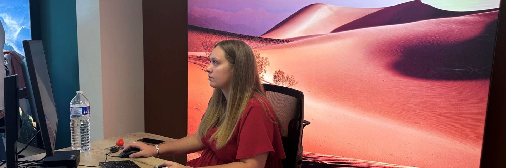 Eine Frau mit langem blonden Haar sitzt an einem Schreibtisch vor einem Computerbildschirm und tippt auf einer Tastatur, neben ihr stehen eine Wasserflasche und Unterlagen; im Hintergrund ist ein großformatiges Wandbild mit rosa-orangefarbenen Sanddünen in Wüstenlandschaft zu sehen.