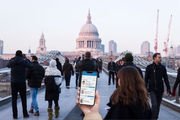 Eine Frau hält ein Smartphone mit einer medizinischen App auf dem Display, während sie über die belebte Millennium Bridge in London in Richtung St. Paul’s Cathedral geht; im Hintergrund sind viele Passanten und die berühmte Kuppel zu sehen