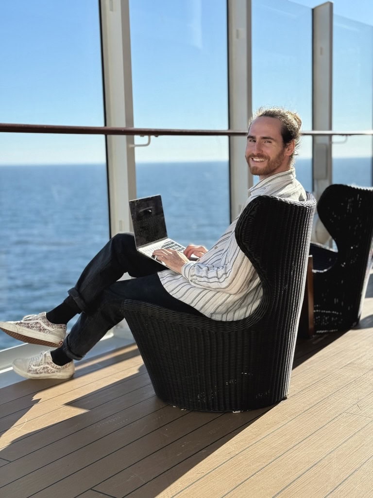 Sales-Coach Ricco Groß, ein Mann Anfang 30, sitzt mit Laptop auf dem Schoß auf einem Korbsessel im Außenbereich eines Kreuzfahrtschiffes, im Hintergrund das Meer.