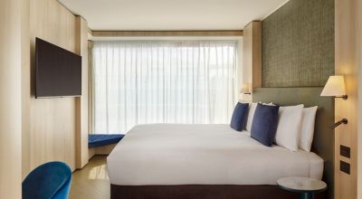 Modernes Hotelzimmer in London mit großem Doppelbett, eleganter Inneneinrichtung und bodentiefem Fenster mit lichtdurchlässigem Vorhang; ein Flachbildfernseher hängt an der Wand und ein blauer Sessel steht am Schreibtisch.