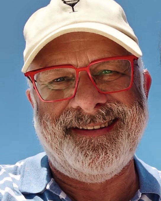 Ein Mann mit weißem Bart und roter Brille und weißem Käppi lacht in die Kamera