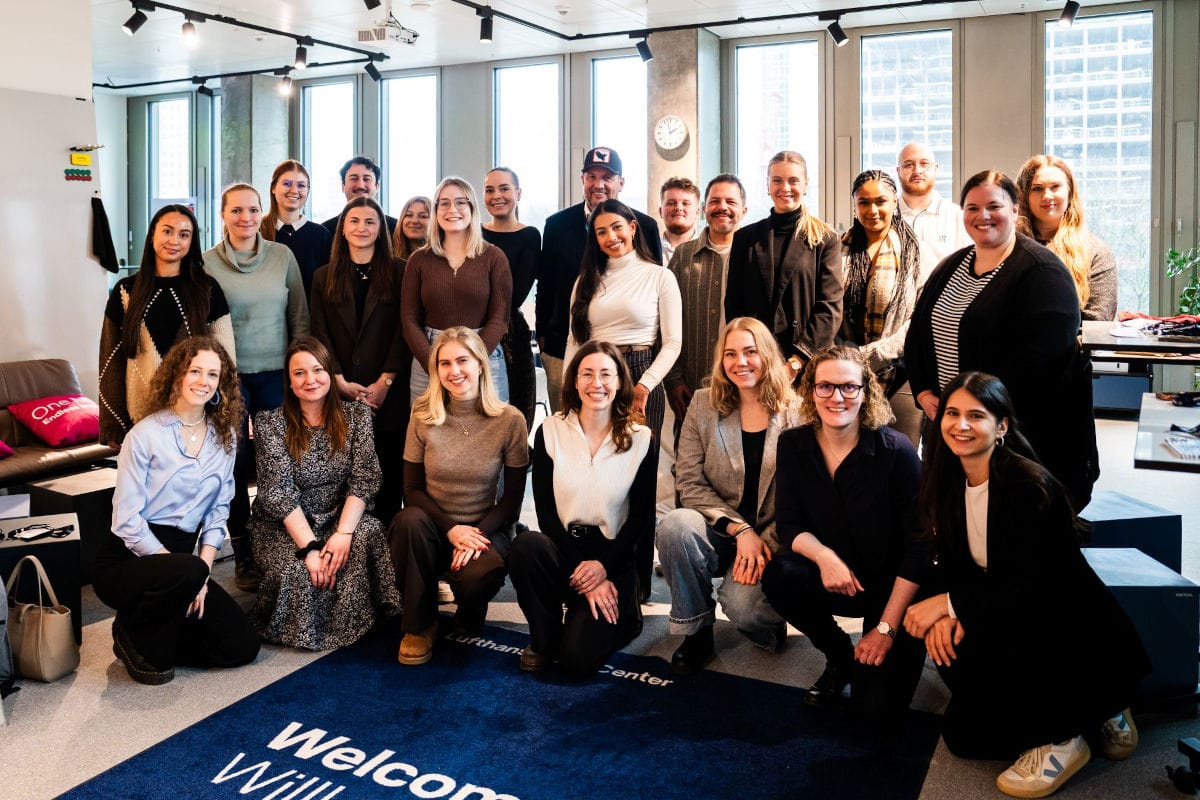 Gruppenfoto von rund 20 Personen in einem modernen, hellen Büro mit großen Fenstern. Das Team steht und kniet in zwei Reihen und lächelt in die Kamera. Im Vordergrund liegt ein blauer Teppich mit der Aufschrift „Welcome Willa“ und „Lufthansa Innovation Center“. Im Hintergrund sind Arbeitsbereiche, eine Uhr an der Wand und Lounge-Möbel zu sehen.