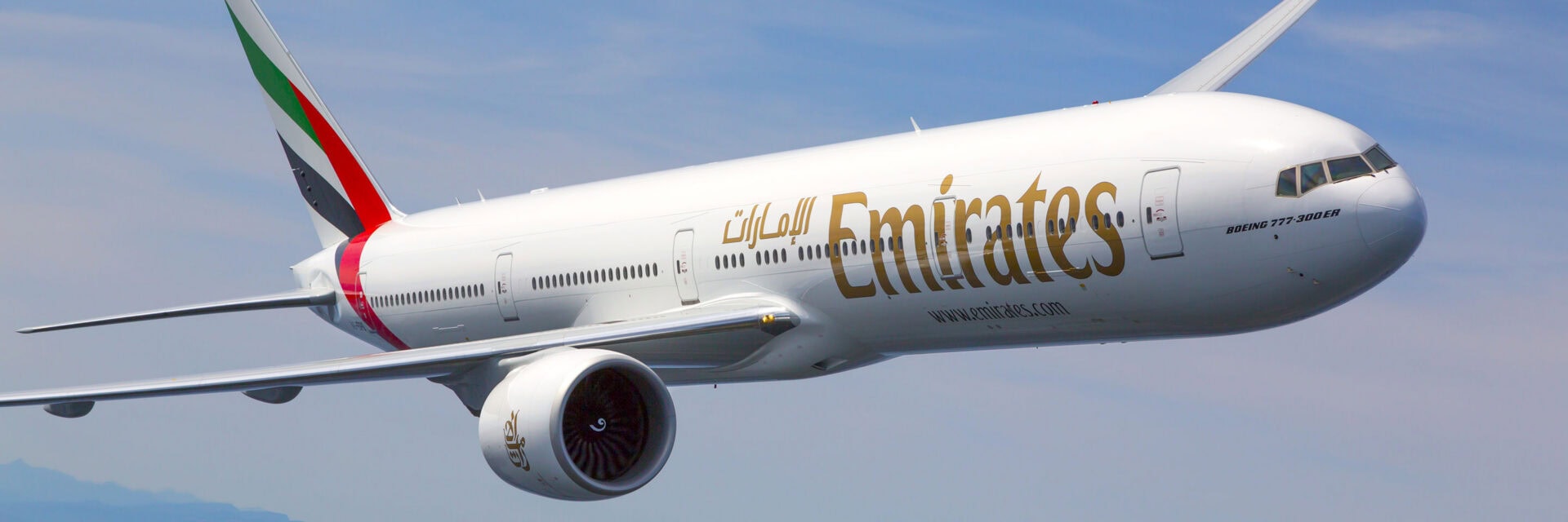 Eine Emirates Boeing 777-300ER fliegt durch den Himmel, das Flugzeug ist in Nahaufnahme zu sehen und hebt sich deutlich vor dem blauen Hintergrund ab. Das auffällige Emirates-Logo und die arabische Schrift sind auf dem weißen Rumpf des Passagierflugzeugs gut sichtbar.