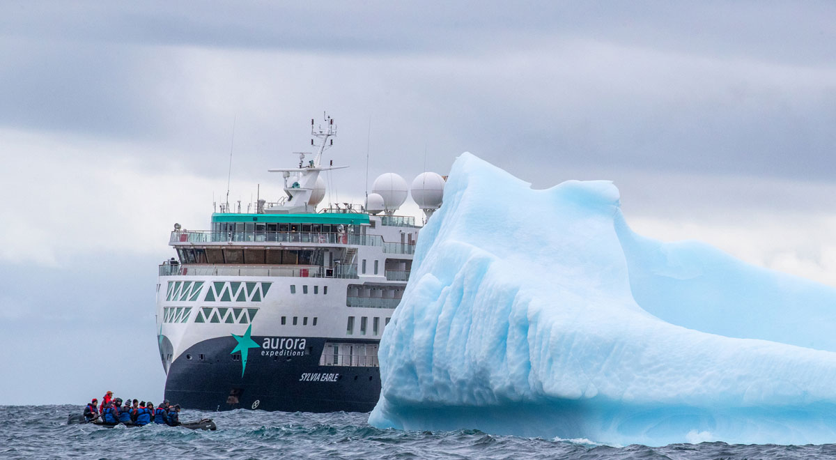 Ein modernes Expeditionsschiff mit dem Namen „Sylvia Earle“ fährt im arktischen Ozean dicht an einem riesigen, hellblauen Eisberg vorbei, während ein kleines Schlauchboot mit Menschen im Vordergrund das Abenteuer in der Arktis sucht. Das Bild zeigt eindrucksvoll die majestätische Natur und die Faszination von Expeditionskreuzfahrten in polaren Regionen.
