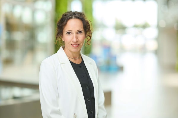 Porträt einer Frau mit hochgesteckten, lockigen braunen Haaren, die lächelnd in die Kamera blickt. Sie trägt ein weißes Sakko über einer dunklen Bluse. Der Hintergrund ist hell, modern und unscharf, mit grünen Elementen, die auf ein zeitgemäßes Innenraum- oder Büroambiente hindeuten.