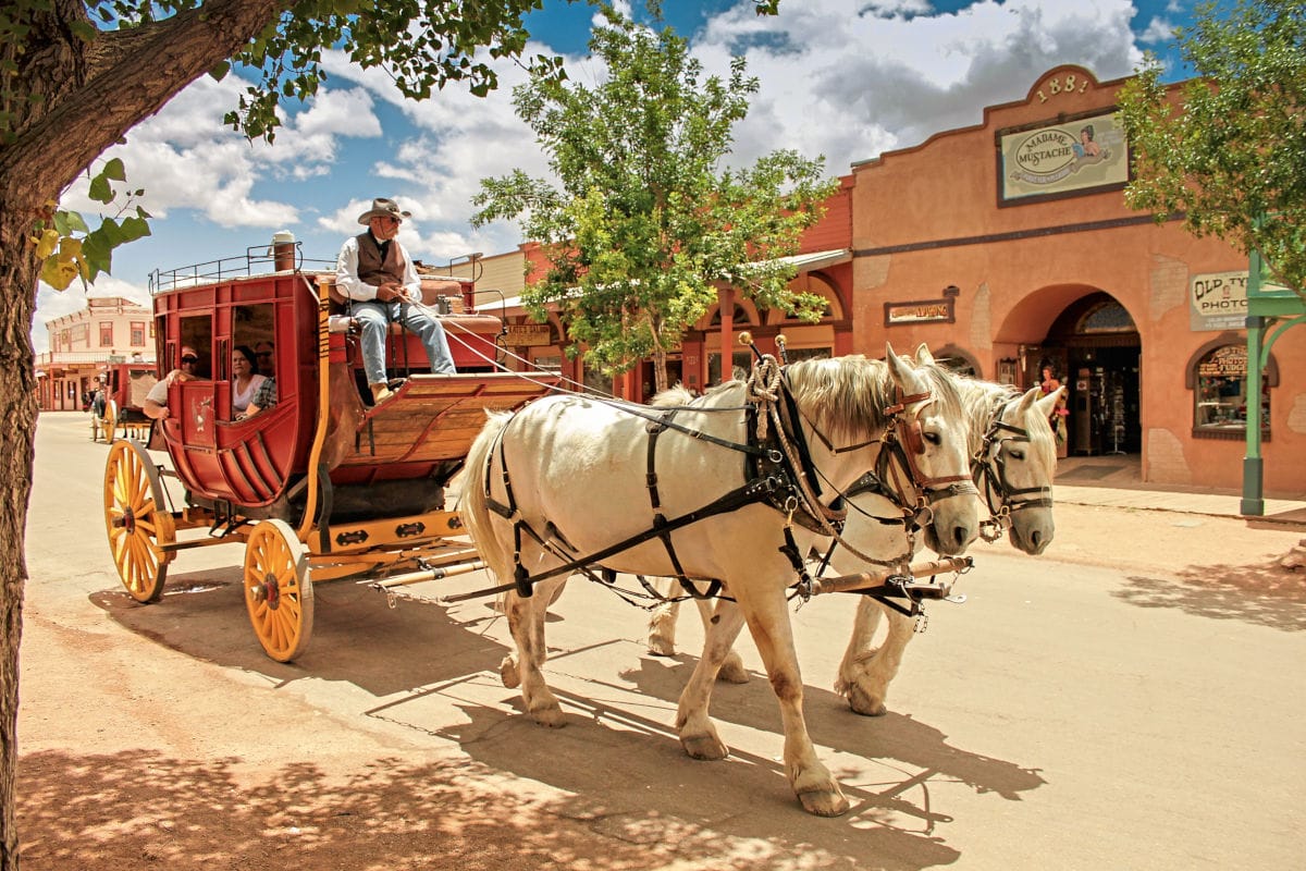 Auf dem Bild fährt eine rote Postkutsche, gezogen von zwei weißen Pferden, durch die historische Westernstadt Tombstone in Arizona. Im Vordergrund sitzen der Kutscher und Passagiere im nostalgischen Old-West-Setting, während im Hintergrund typische Westerngebäude zu sehen sind.