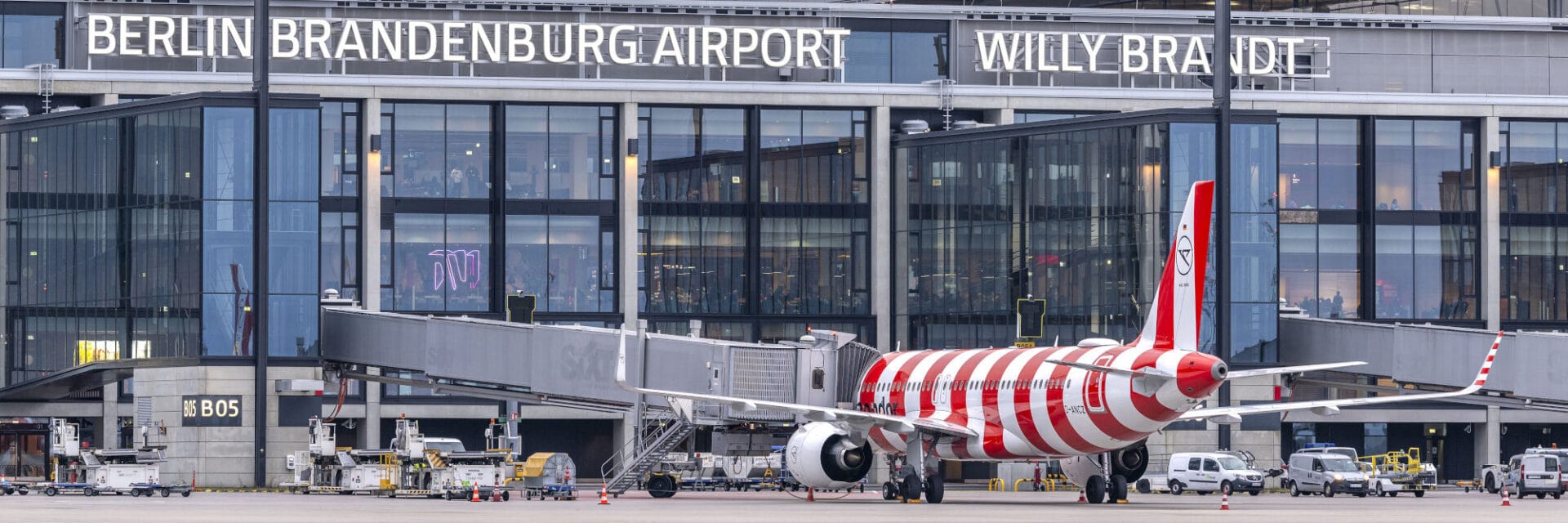 Ein Condor-Flugzeug mit auffälligen rot-weißen Streifen steht am Terminal des Berliner Flughafens, direkt angeschlossen an eine Fluggastbrücke. Im Hintergrund ist die moderne Glasfassade des Flughafengebäudes mit gut sichtbarem Schriftzug zu sehen. Auf dem Vorfeld befinden sich Bodenfahrzeuge und Gepäckwagen, die den Betrieb am Flughafen Berlin unterstützen.