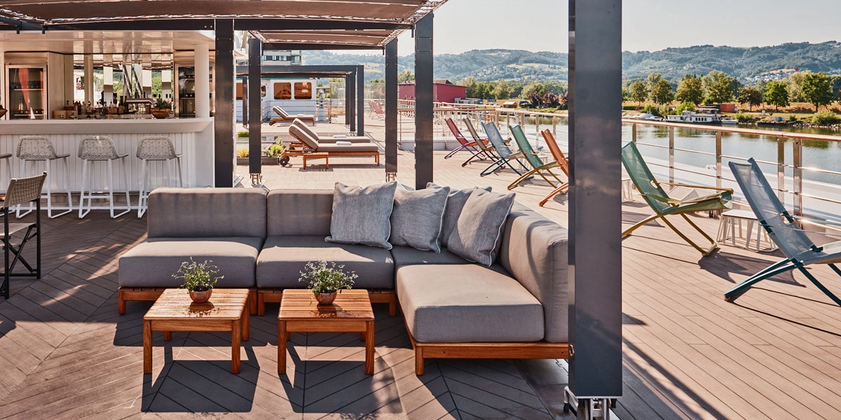 Luxuriöse Lounge-Ecke auf dem Sonnendeck eines Flusskreuzfahrtschiffs mit modernen Sofas, Holztischen und Blick auf den Fluss sowie Liegestühlen im Hintergrund. Die Szene vermittelt entspannte Urlaubsatmosphäre auf einer Golfkreuzfahrt.