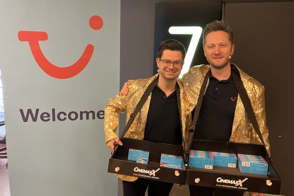 Zwei lächelnde Männer in goldenen Jacken stehen nebeneinander und halten Snack-Tabletts vor einem CinemaxX-Hintergrund; links ist ein „Welcome“-Banner mit Logo zu sehen.