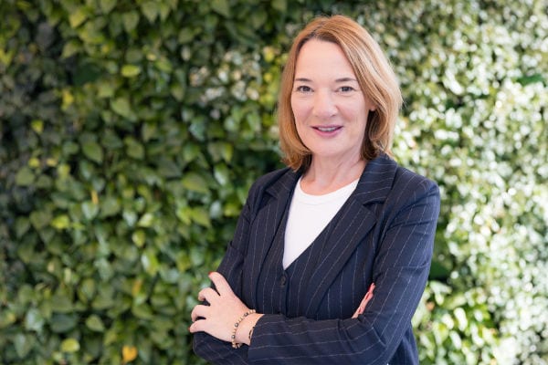 Porträt einer lächelnden Frau mit schulterlangem, hellbraunem Haar, die vor einer grünen Pflanzenwand steht und mit verschränkten Armen einen dunklen Blazer über einem weißen Oberteil trägt.