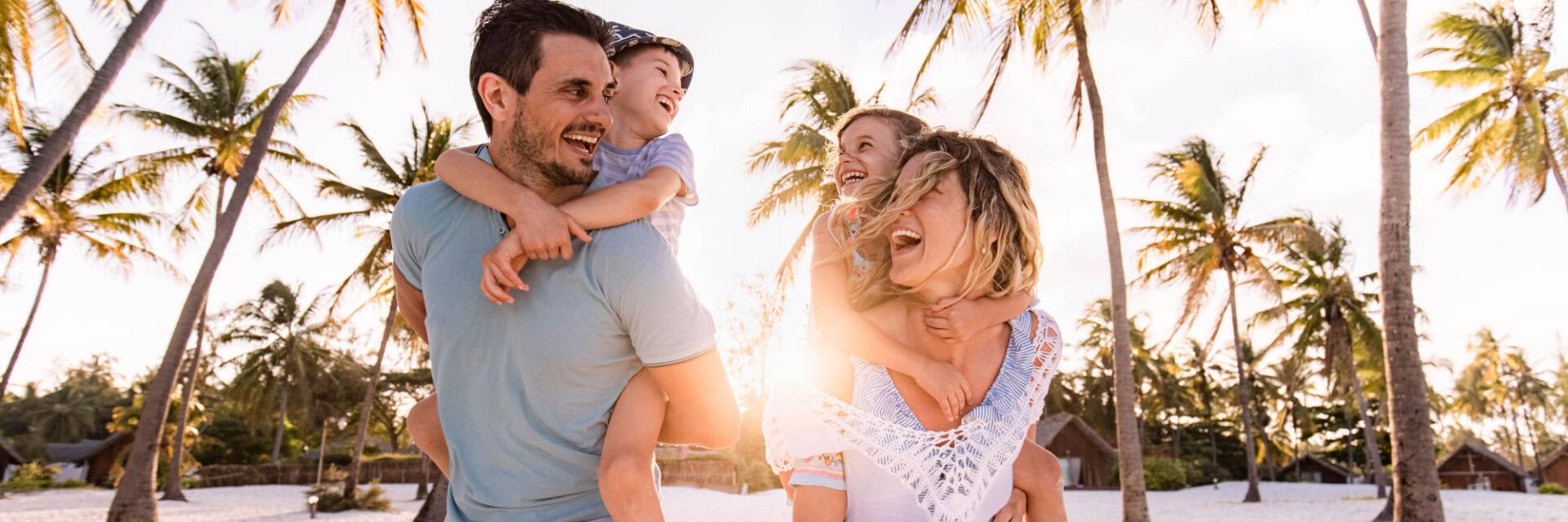 Fröhliche Familie am Strand: Zwei Erwachsene tragen jeweils ein Kind auf dem Rücken, alle lachen, im Hintergrund Palmen und warmes Abendlicht.