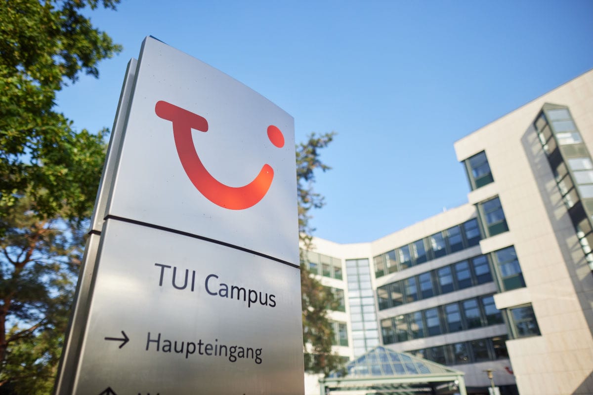 Schild mit rotem TUI-Logo und der Aufschrift „TUI Campus Haupteingang“ vor modernen Bürogebäuden, umgeben von Bäumen unter blauem Himmel.