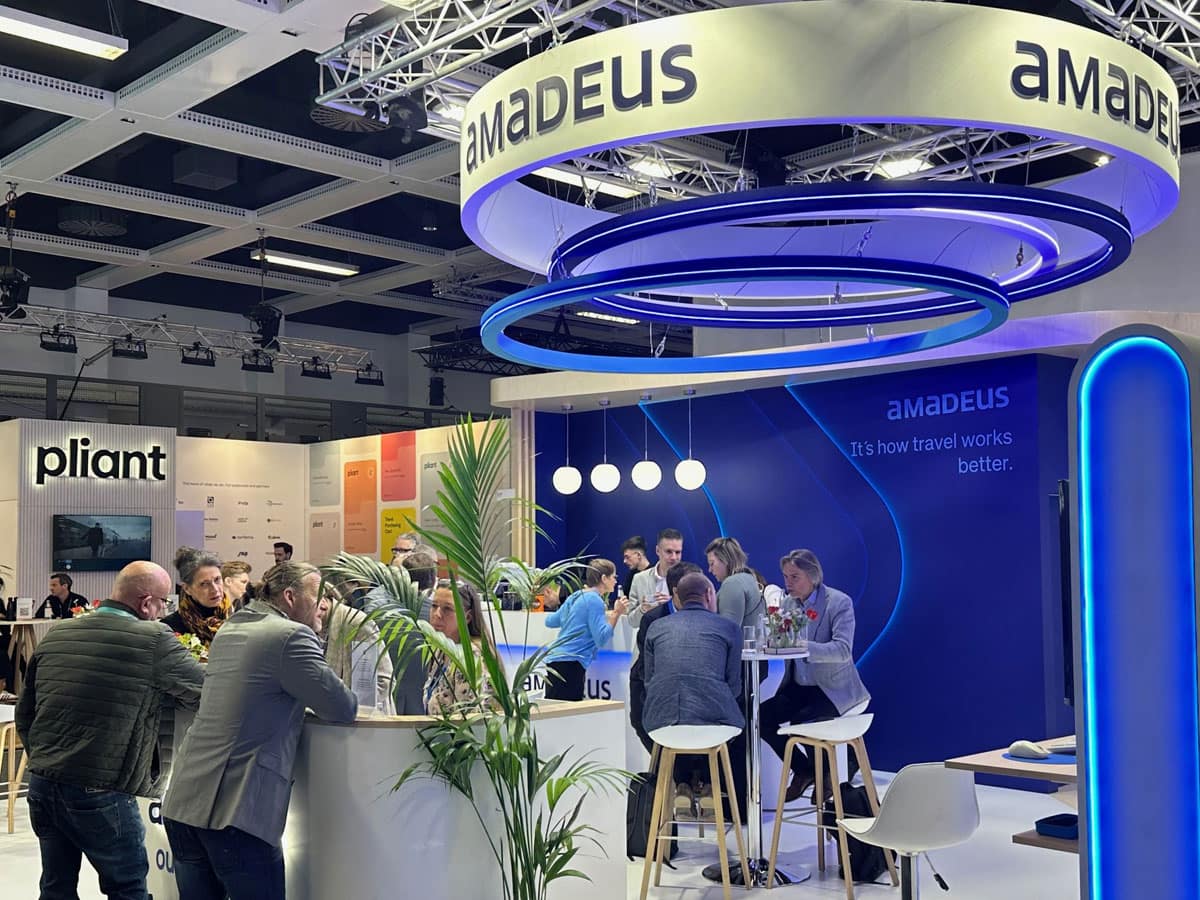 Messestand von Amadeus auf einer Messehalle mit blau-weißer Beleuchtung, runden Deckenringen, Stehtischen und mehreren Menschen im Gespräch; im Hintergrund weitere Stände, unter anderem mit dem Schriftzug „pliant“.