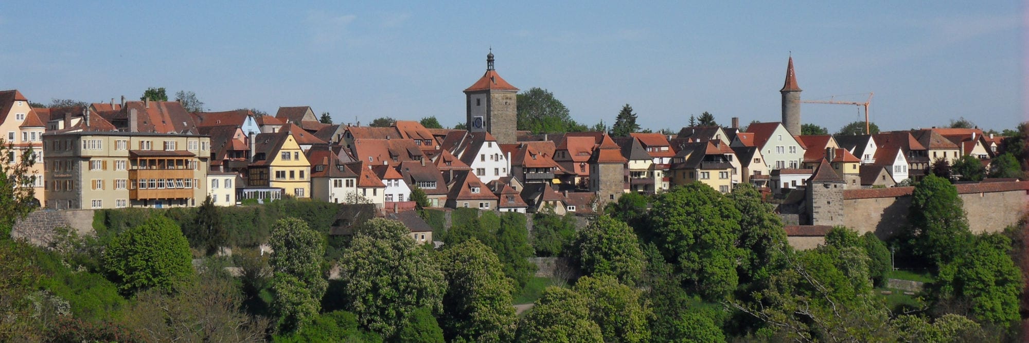 Blick auf eine historische Altstadt mit roten Ziegeldächern, Fachwerkhäusern und steinernen Türmen, umgeben von Stadtmauern und grünen Bäumen unter blauem Himmel.