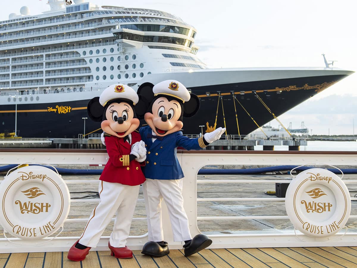 Vor dem imposanten Kreuzfahrtschiff Disney Wish stehen zwei Disney-Charaktere in Kapitänsuniform und winken herzlich den Passagieren zu. Das Foto vermittelt die Magie des Disney Cruise Line Erlebnisses am Hafen. Die weißen Rettungsringe mit dem goldenen Disney Wish Logo unterstreichen das maritime Thema.