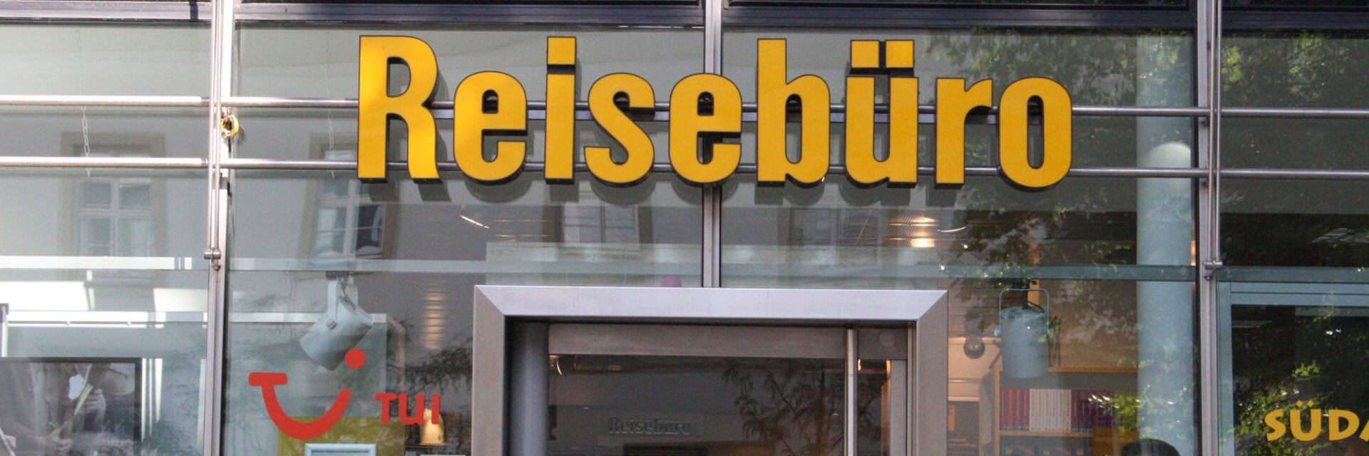 Auf dem Foto ist die Glasfassade eines Reisebüros in der Innenstadt zu sehen, mit großer gelber Aufschrift „Reisebüro“ und dem TUI-Logo. Das Schaufenster spiegelt die Umgebung wider und gibt einen Einblick in das Innere des Reisebüros.