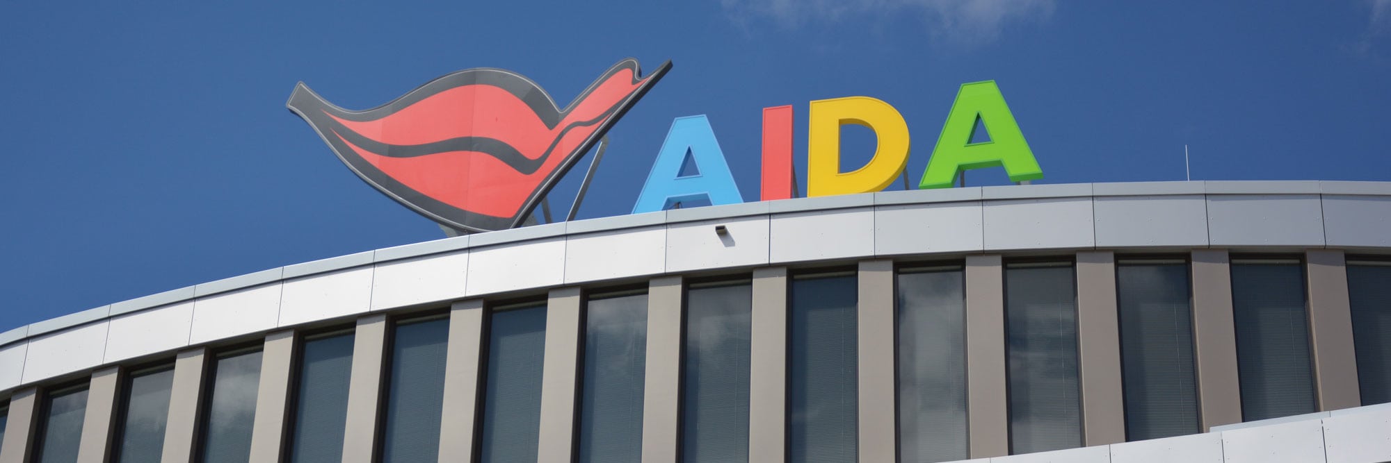 Das Foto zeigt die moderne Fassade der AIDA Zentrale mit großem, farbigem AIDA Logo und stilisiertem roten Kussmund auf dem Dach vor blauem Himmel.