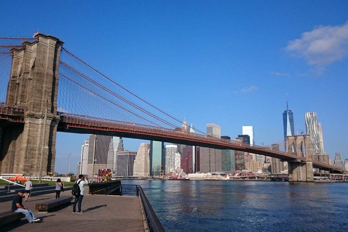 Die Brooklyn Bridge spannt sich über den East River vor der Skyline von Manhattan mit dem One World Trade Center im Hintergrund. Im Vordergrund stehen und sitzen Menschen auf einer Uferpromenade unter blauem Himmel.
