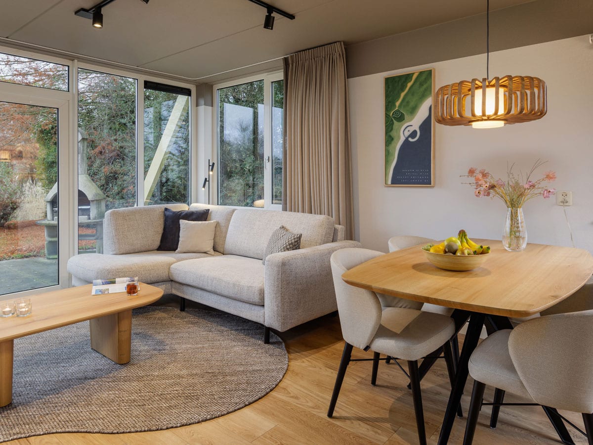 Helles, modern eingerichtetes Wohnzimmer mit großer Ecksofa in Beige, rundem Teppich und Holzcouchtisch, daneben Essbereich mit Holztisch und gepolsterten Stühlen, großen Fenstern mit Blick ins Grüne und einer Hängelampe aus Holz.