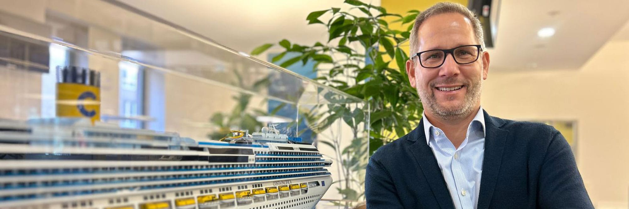 Ein Manager steht neben einem großen Modell eines Kreuzfahrtschiffes der Marke Costa, im Hintergrund sind grüne Pflanzen und ein modernes Büro zu sehen. Das Bild zeigt deutliche Details des Schiffsmodells, wie die gelben Rettungsboote und den charakteristischen Schornstein mit Logo.