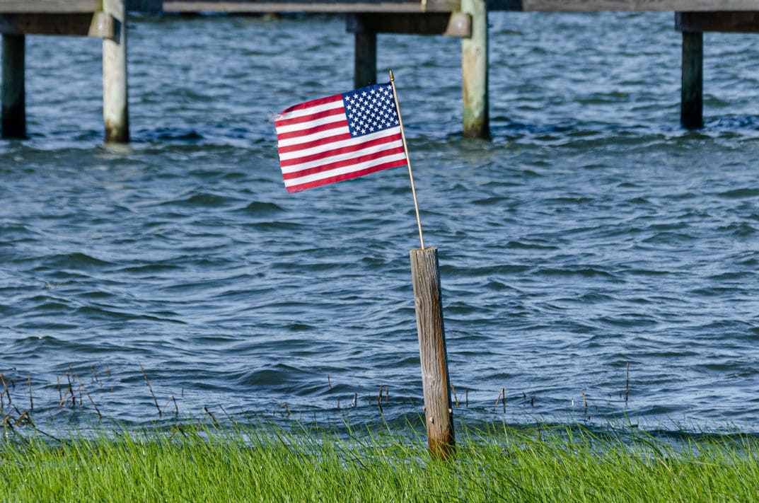 Die US-Flagge ist an einem schrägen Holzpfosten befestigt und steht im Vordergrund vor einem Gewässer mit sanften Wellen und einem Bootssteg.
