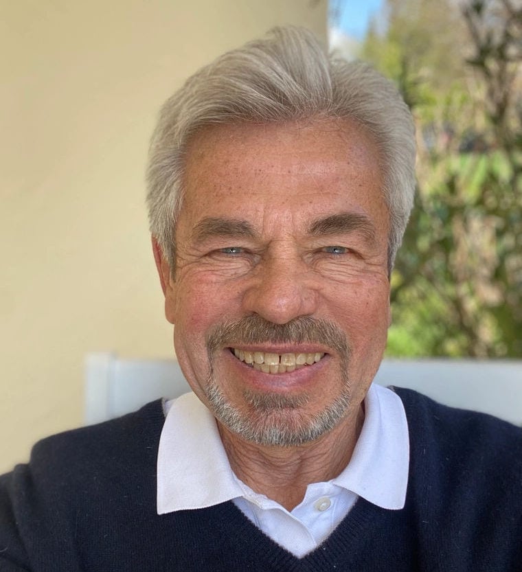 Ein älterer Mann mit grauem Haar sitzt draußen vor einer hellen Wand, trägt ein weißes Hemd und einen dunkelblauen Pullover; im Hintergrund sind Bäume und Sträucher zu sehen.