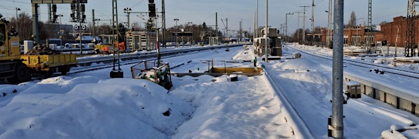 Auf dem Foto sind schneebedeckte Bahngleise an einem Bahnhof der Deutschen Bahn zu sehen, mit Bauarbeiten und Arbeitsfahrzeugen an den Gleisen. Die winterliche Landschaft zeigt, wie der Zugverkehr im Winter durch Schnee und Kälte beeinträchtigt werden kann.