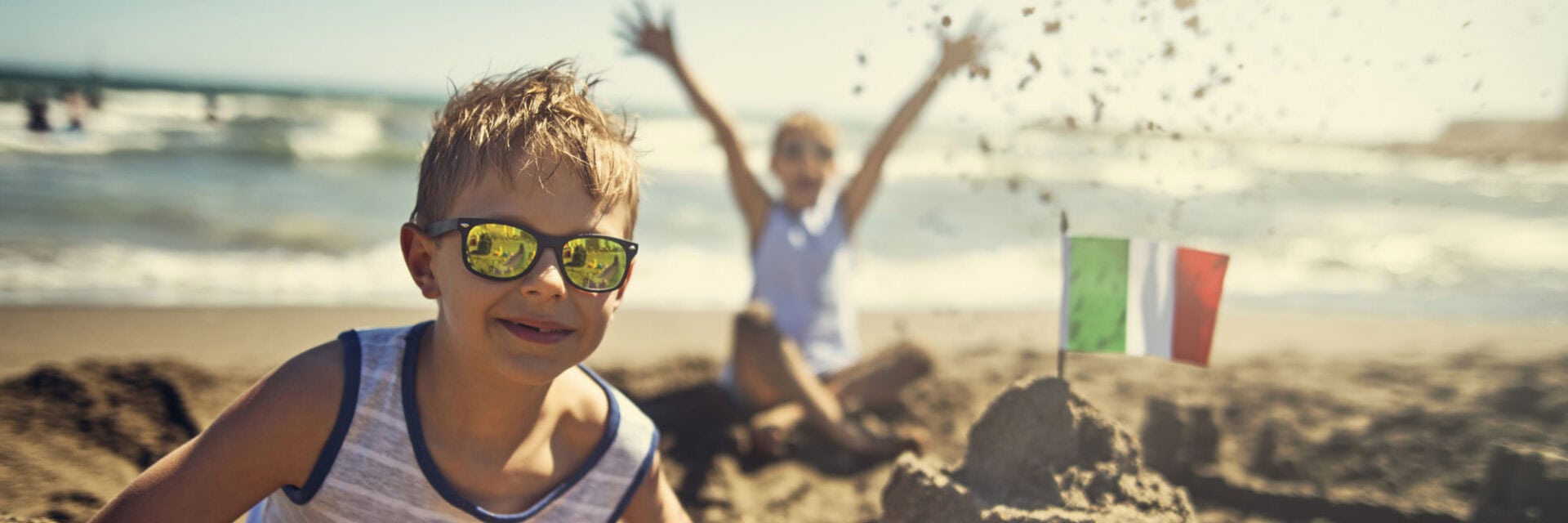 Das Bild zeigt zwei Kinder am italienischen Strand beim Bau einer Sandburg, die stolz mit einer kleinen Italien-Flagge dekoriert ist, während die Sonne scheint und das Meer im Hintergrund glitzert – perfekte Szene für Italien Urlaub mit Kindern und Strandspaß.