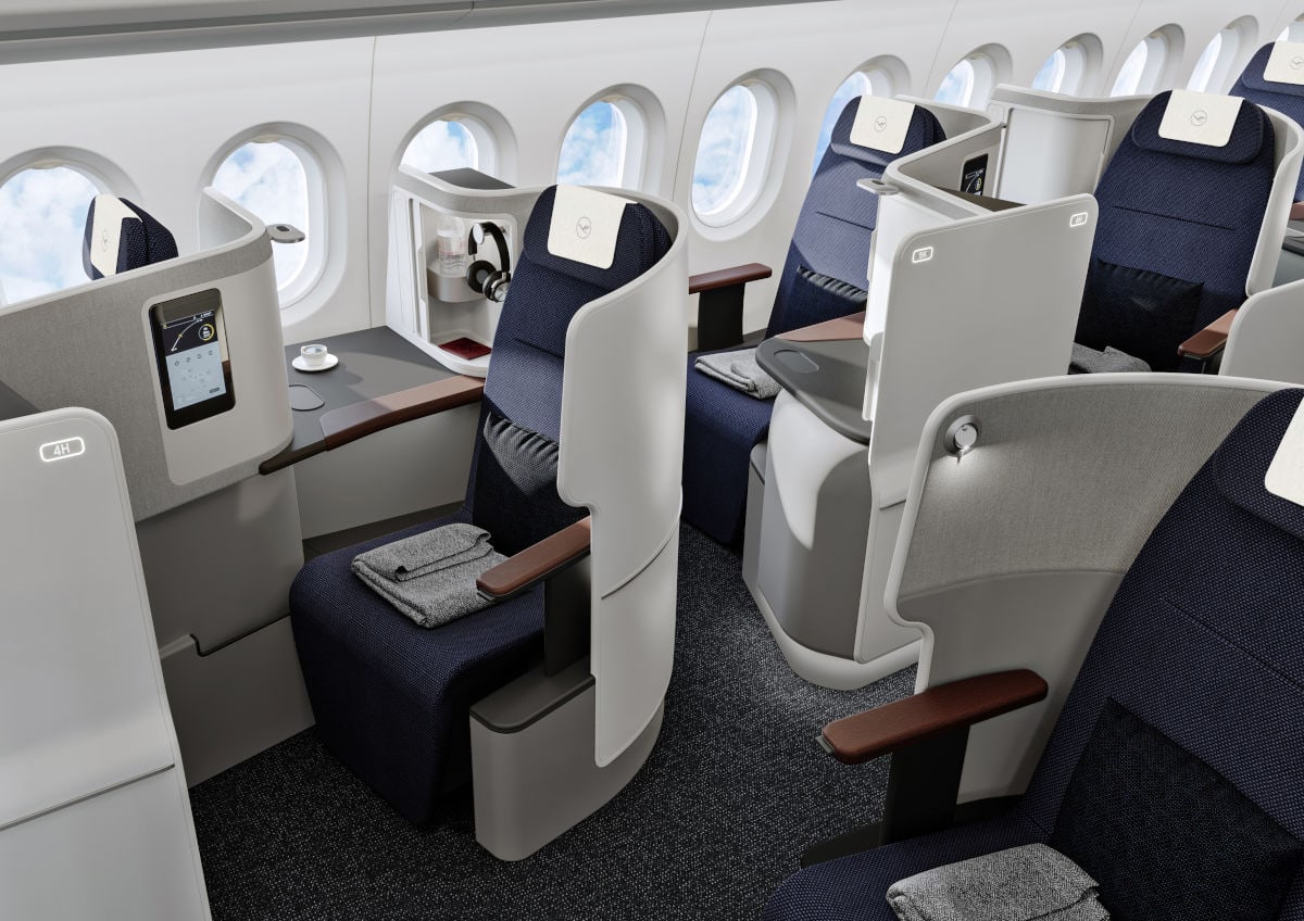 Im Bild sind moderne Lufthansa Business Class Sitze in einem Flugzeug zu sehen, die mit komfortablen Polstern, großzügigem Abstand und persönlichen Ablagen ausgestattet sind. Die Sitze bieten Privatsphäre, eigene Bildschirme sowie Fenster mit Blick auf den Himmel.